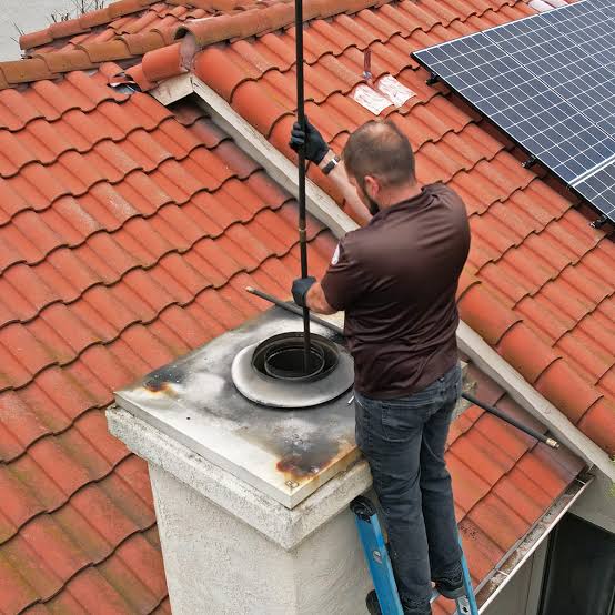 Technician cleaning a round flue on a stucco chimney surrounded by red clay roof tiles